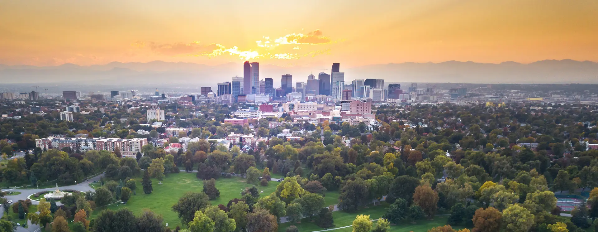 Aerial view of a city skyline at sunset, with vibrant orange and pink hues. Lush green parks in the foreground and mountains silhouetted in the distance