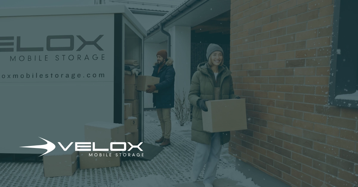 A man and woman wearing winter coats carry moving boxes from a brick house into a weather-sealed Velox Mobile Storage container during a snowy Utah day.
