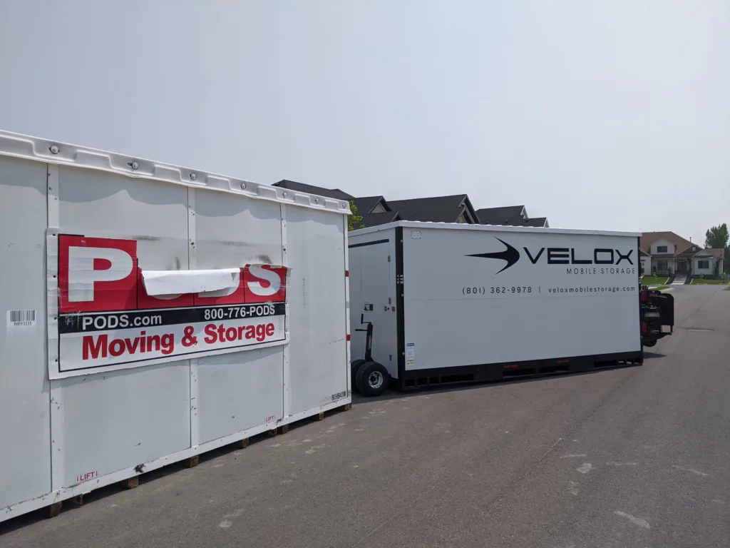 A PODS container and a Velox Mobile Storage container parked side by side on a Utah street, showing the Velox unit's steel body and black base frame