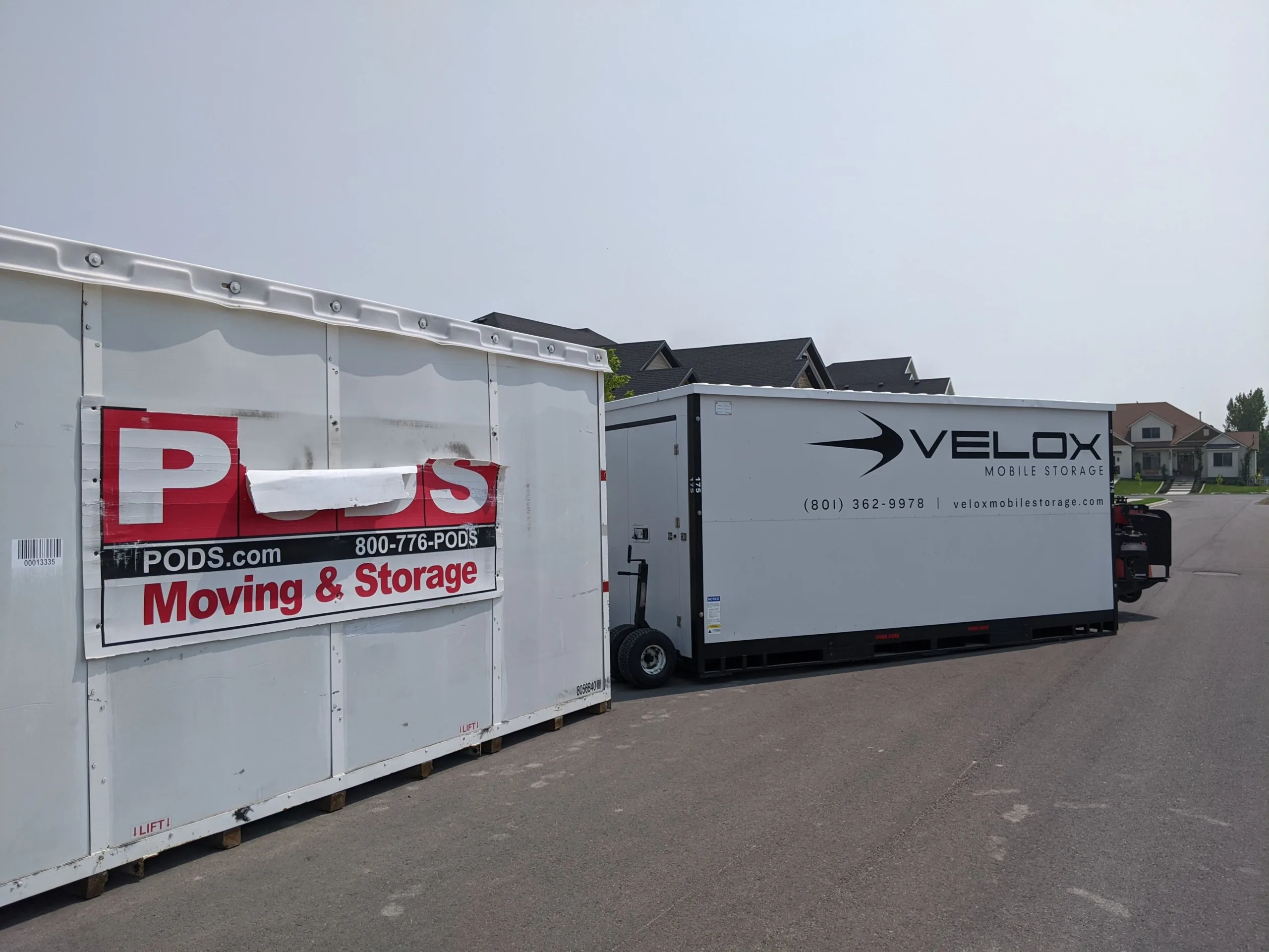 A PODS container and a Velox Mobile Storage container parked side by side on a Utah street, showing the Velox unit's steel body and black base frame