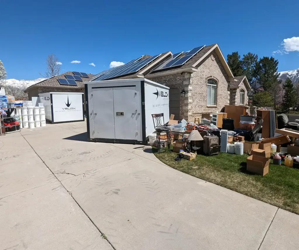 Two Velox portable storage containers on a driveway in Highland, Utah, with household belongings set out on the lawn during a whole-home project